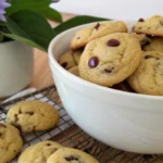 A white bowl filled with golden little chocolate chip cookies, with a purple flower on a cooling rack.