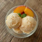 Two scoops of delicious homemade peach ice cream, garnished with fresh peach slices and mint, in a clear glass bowl.