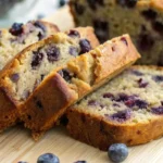 Sliced blueberry banana bread on a wooden board with fresh blueberries. Homemade quick bread is ready to serve.