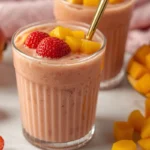 Two vibrant strawberry mango smoothies in glasses, garnished with fresh fruit, surrounded by whole mangoes and strawberries.