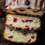 Slices of glazed cranberry orange loaf on a dark surface, with fresh cranberries and orange zest.