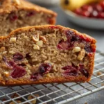 A thick slice of moist banana cranberry bread with vibrant cranberries and nuts on a wire rack.