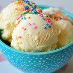 Three scoops of cake batter ice cream with rainbow sprinkles in a blue bowl, waffle cone behind.