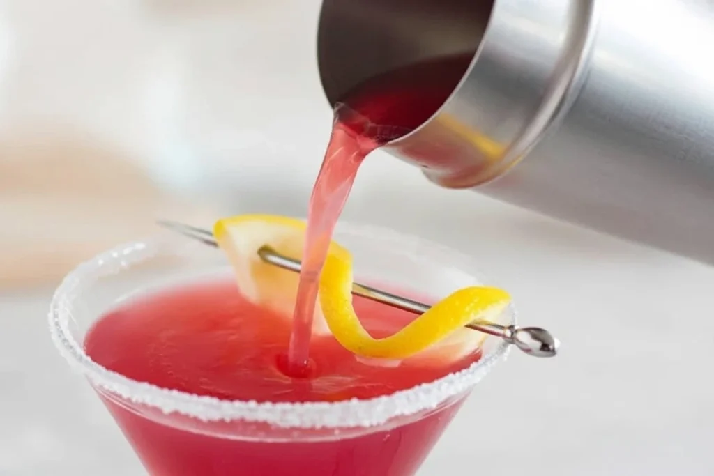 Cranberry lemon drop cocktail being poured into a sugared martini glass with a lemon twist.