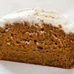 Close-up of an angled slice of pumpkin zucchini bread on parchment, showing its moist crumb with green zucchini and thick white frosting.